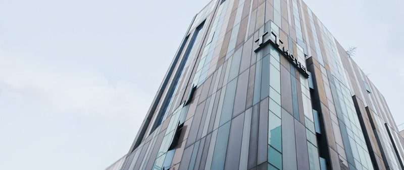 트리아호텔 건물 외관 — 회색·청록 세로 패널 외벽 모서리의 TRIA HOTEL 간판, 흐린 하늘