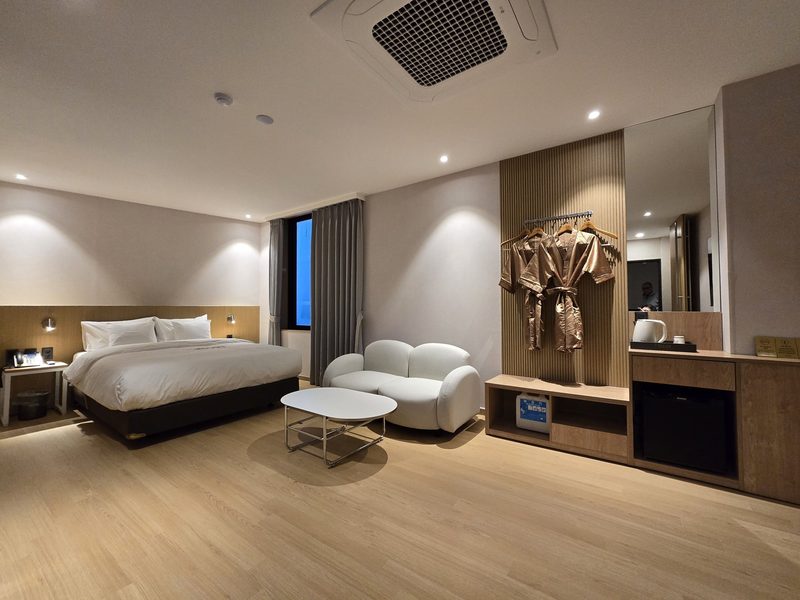 디럭스 더블룸 — 침대와 흰 소파, 가운 행거와 미니 냉장고 수납장이 보이는 객실 전체
